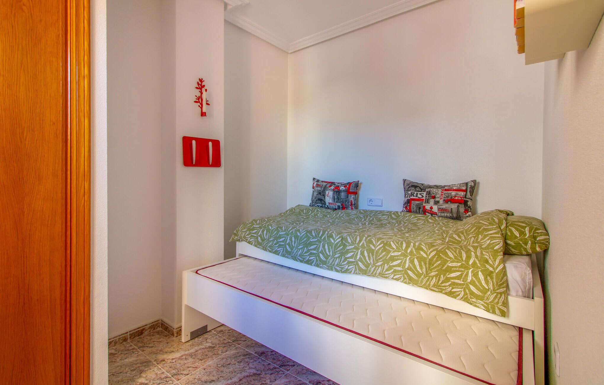 A small, single-bed room with a green patterned bedspread, two pillows with a cityscape design, and a red wall-mounted light switch.