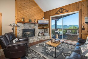 A cozy living room with a fireplace, leather sofas, a stone wall, and a large sliding glass door leading to an outdoor deck. - Pet-Friendly Retreat with Hot Tub, Fire Pit & Mountain Views (Banner Elk)
