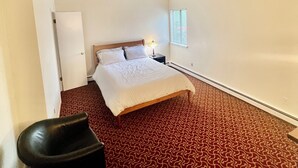 A hotel room with a large bed, a small bedside table with a lamp, a window with blinds, and a patterned carpet. - 🤣🏡 "Welcome to our basement suite, where humor and professionalism coexist! (Peekskill)