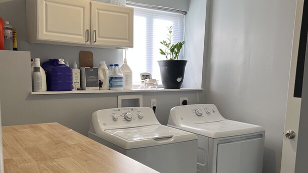 A laundry room with two washing machines, a wooden table, and a window with blinds. - 🤣🏡 "Welcome to our basement suite, where humor and professionalism coexist! (Peekskill)
