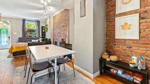 A dining area with a brick wall, a white table, and chairs. There is a bookshelf with books and decorative items.
