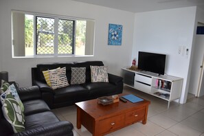 A living room with a black leather sofa, a wooden coffee table, a TV on a white stand, and a bookshelf. - Tupe's Beach House (Vaimaanga)