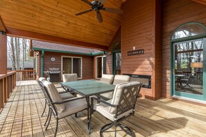 Une terrasse extérieure en bois, avec une table et des chaises pour les repas, un ventilateur de plafond et une vue sur une maison dotée d’une cheminée.