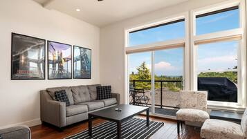 A modern living room with a gray sofa, a black coffee table, and a patterned armchair. There are large windows offering a view of the outdoors.