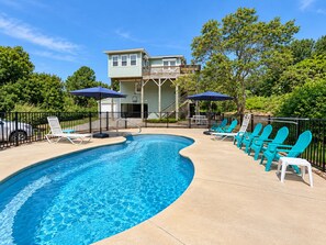 A swimming pool with lounge chairs and umbrellas, surrounded by a fence and trees.