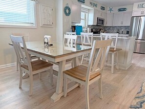 A dining area with a wooden table and chairs, a kitchen in the background, and a light-colored floor.