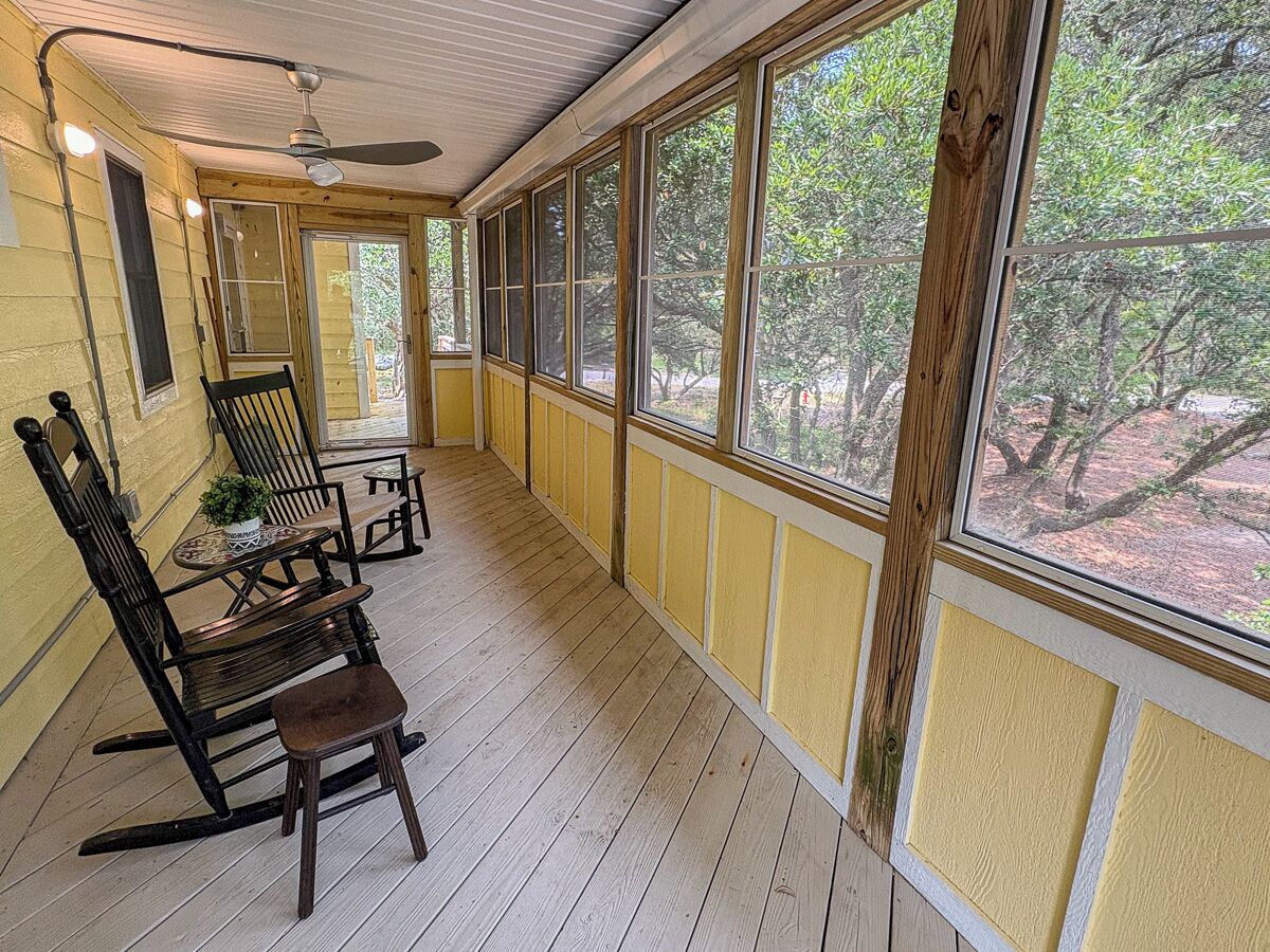 A covered porch with wooden flooring, yellow wainscoting, and large windows offering a view of trees.