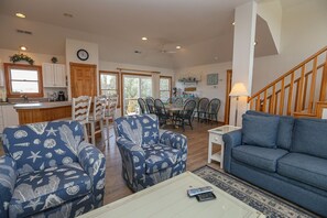 A spacious living room with a blue sofa, patterned armchairs, and a dining area with a wooden table and chairs. A kitchen with white cabinets and a wooden countertop is visible in the background. - Duckaway Home - Unwind in this 5 BR oceanside home in Duck w/Private Pool! (Duck)