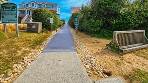 A private oceanfront walkway with a wooden boardwalk, surrounded by houses and dense greenery. - Duckaway Home - Unwind in this 5 BR oceanside home in Duck w/Private Pool! (Duck)