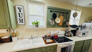 A kitchen with green cabinets, a white sink, a stove, and a microwave. There is a window with blinds, a plant on the windowsill, and a sign that says "YUM". - Green Cottage Farm Stay & Food (Gardner)