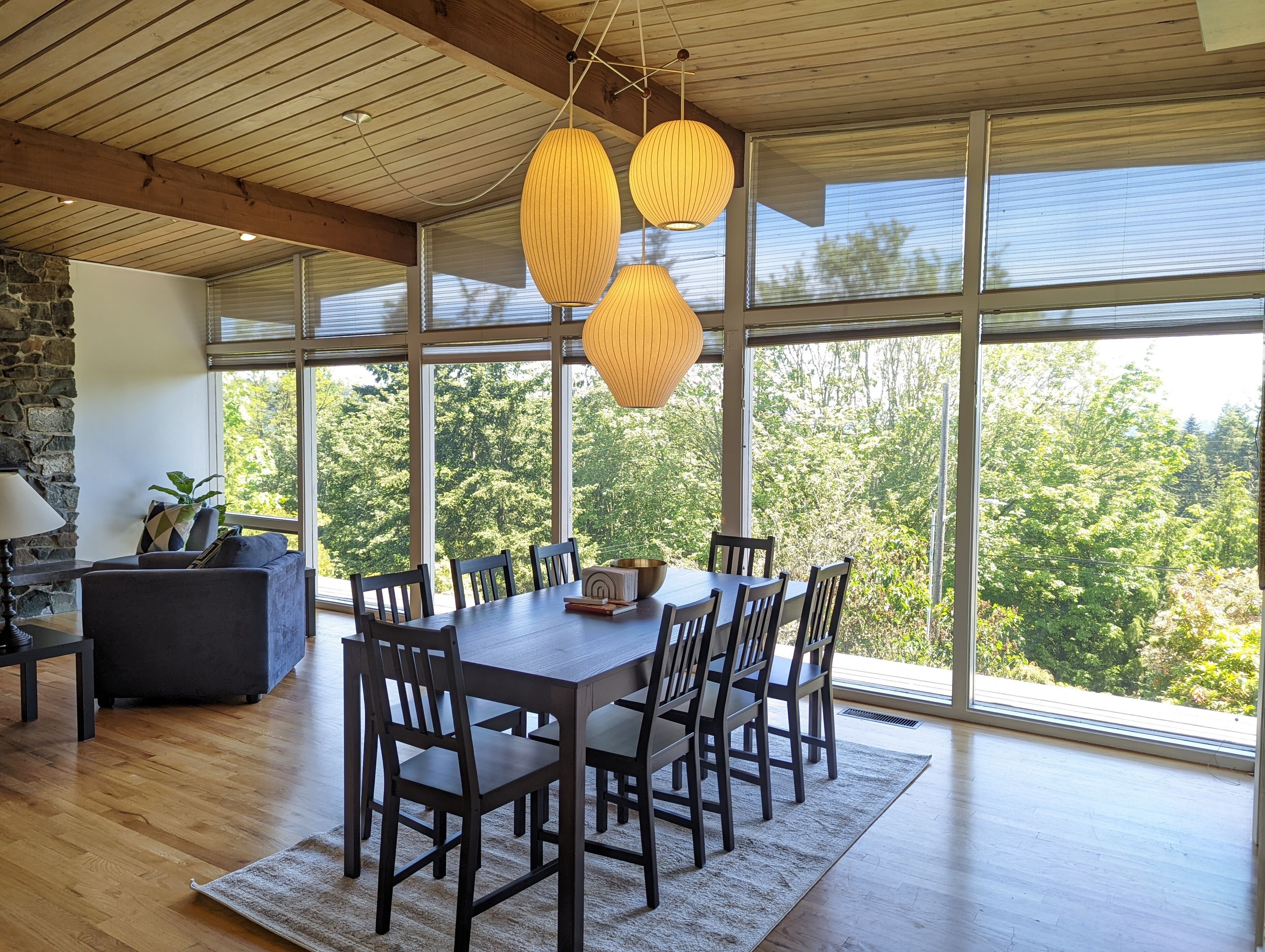 A dining area with a large wooden table and chairs, surrounded by large windows offering a view of trees.