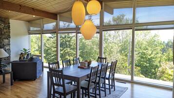 A dining area with a large wooden table and chairs, surrounded by large windows offering a view of trees.