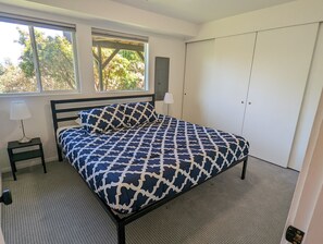 Ein Schlafzimmer mit einem Bett, einem Nachttisch mit Lampe, einem Kleiderschrank und einem Fenster mit Blick auf Bäume.