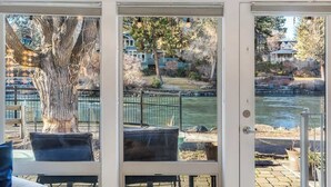A room with a large window overlooking a river, a tree, and a view of houses. - Riverfront Cottage - Steam Room & Pet Friendly! (Bend)