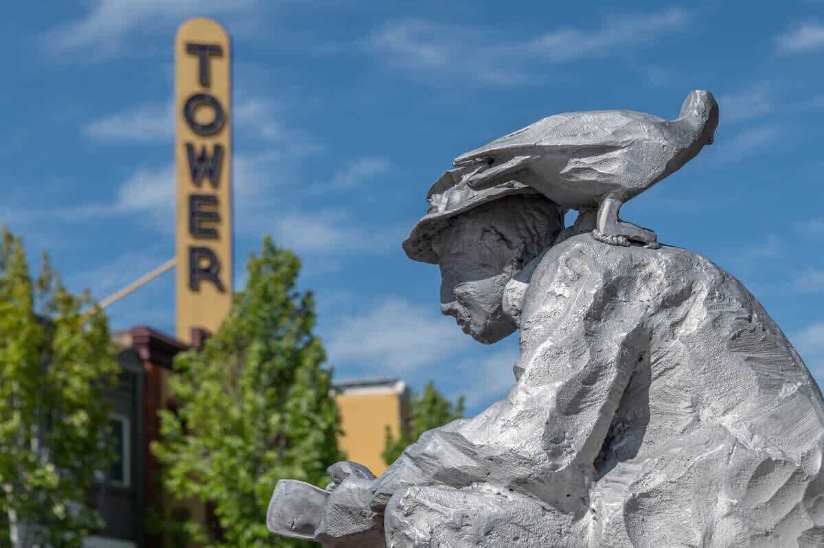 A sculpture of a person with a bird on their shoulder, a sign with the word 'TOWER' in the background, and trees.