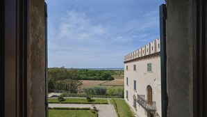 Blick durch ein offenes Fenster auf ein Gebäude mit Garten und eine ferne Landschaft.