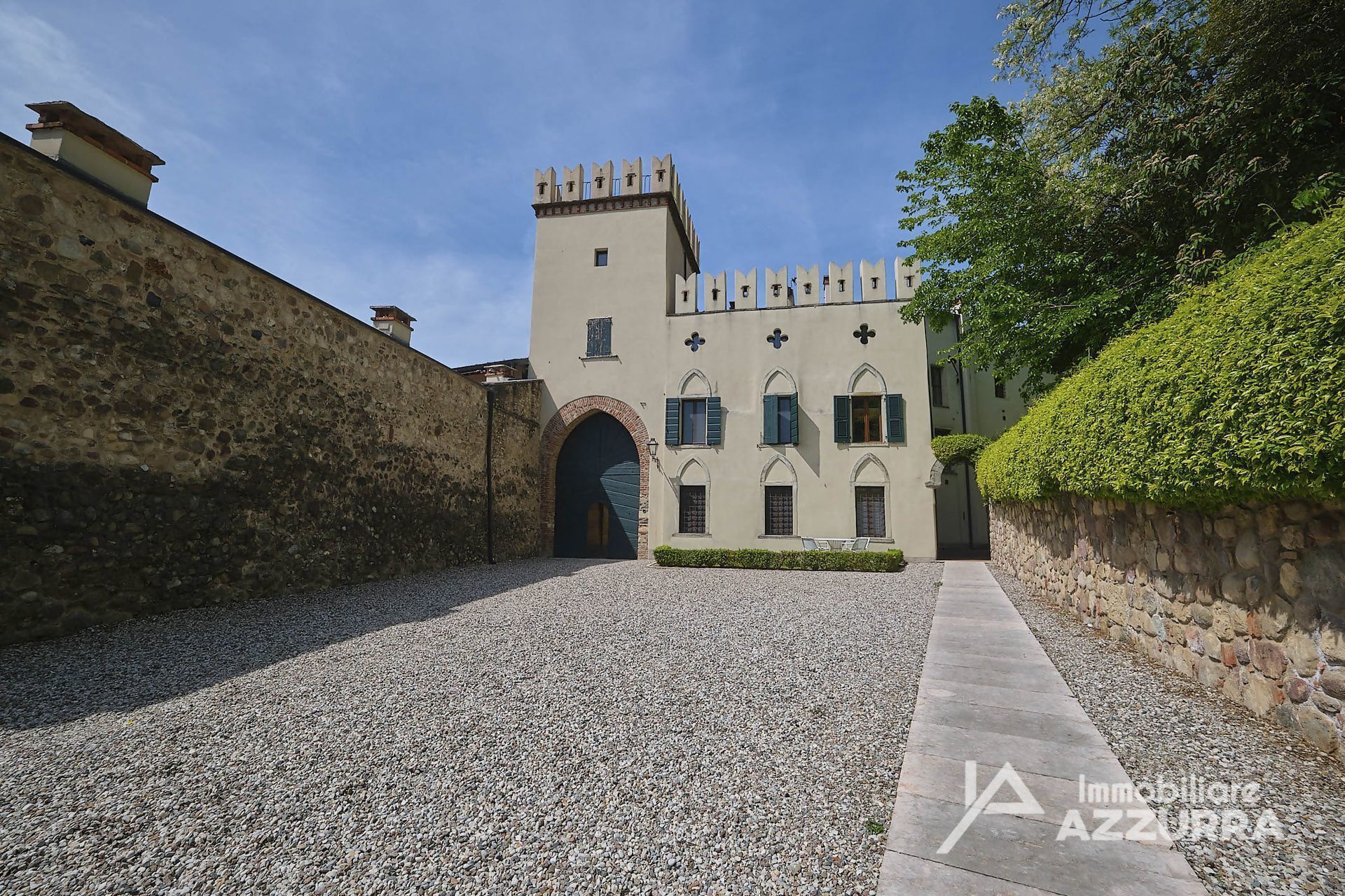 A historic building with a central arched entrance, a tower with battlements, and a gravel courtyard.