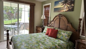 Une chambre avec un grand lit, une tête de lit en bois, un tableau représentant une scène de plage et une vue sur un patio aménagé avec du mobilier d’extérieur.