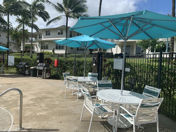 An outdoor patio with white metal tables and chairs, blue umbrellas, and a grill area.