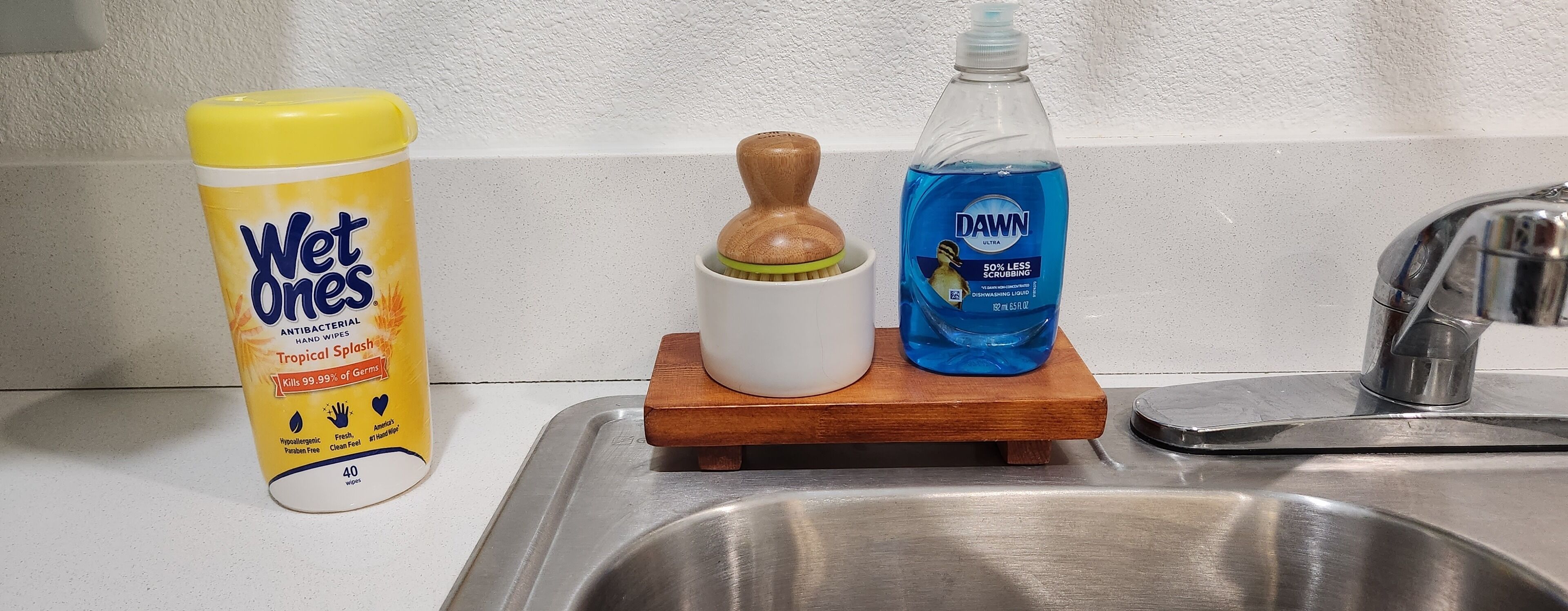 A kitchen sink with a yellow bottle of Wet Ones hand wipes, a wooden brush, and a blue Dawn dishwashing liquid bottle on a wooden tray.