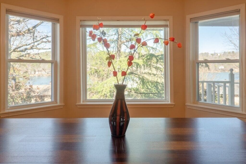 Une pièce avec une table en bois et un vase rempli de fleurs rouges, placé devant une fenêtre donnant sur des arbres et une étendue d’eau.