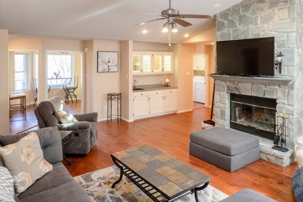 A spacious living room with a stone fireplace, a flat-screen TV, and a dining area with a kitchen in the background.