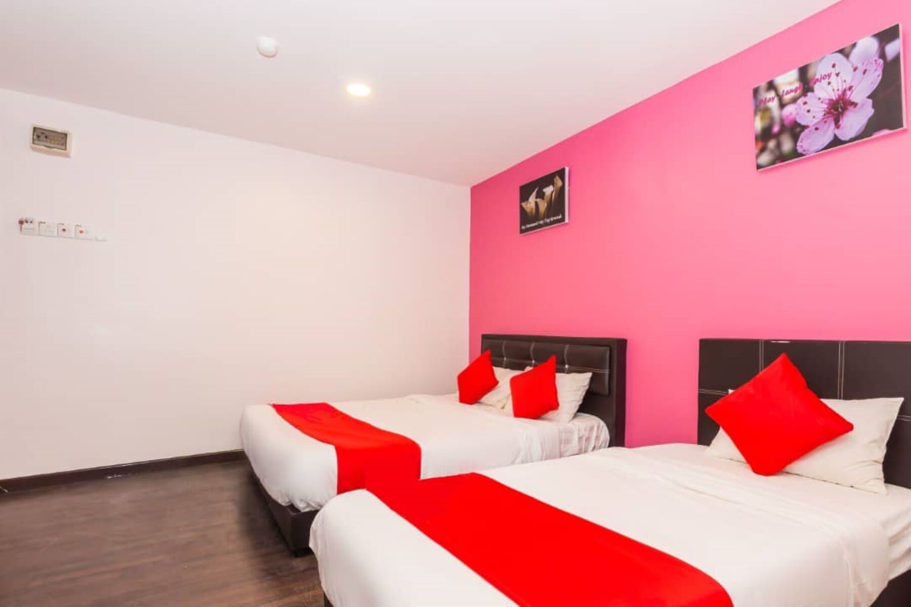 A hotel room with two beds, each with white bedding and red accents, a pink wall, and a framed picture above the beds.