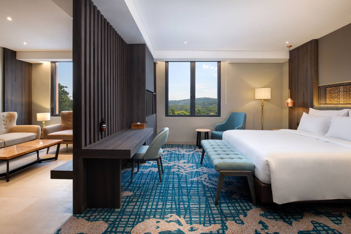 Een hotelkamer met een groot bed, een bureau, een stoel en een bank. Er is uitzicht op het omliggende landschap door een groot raam.