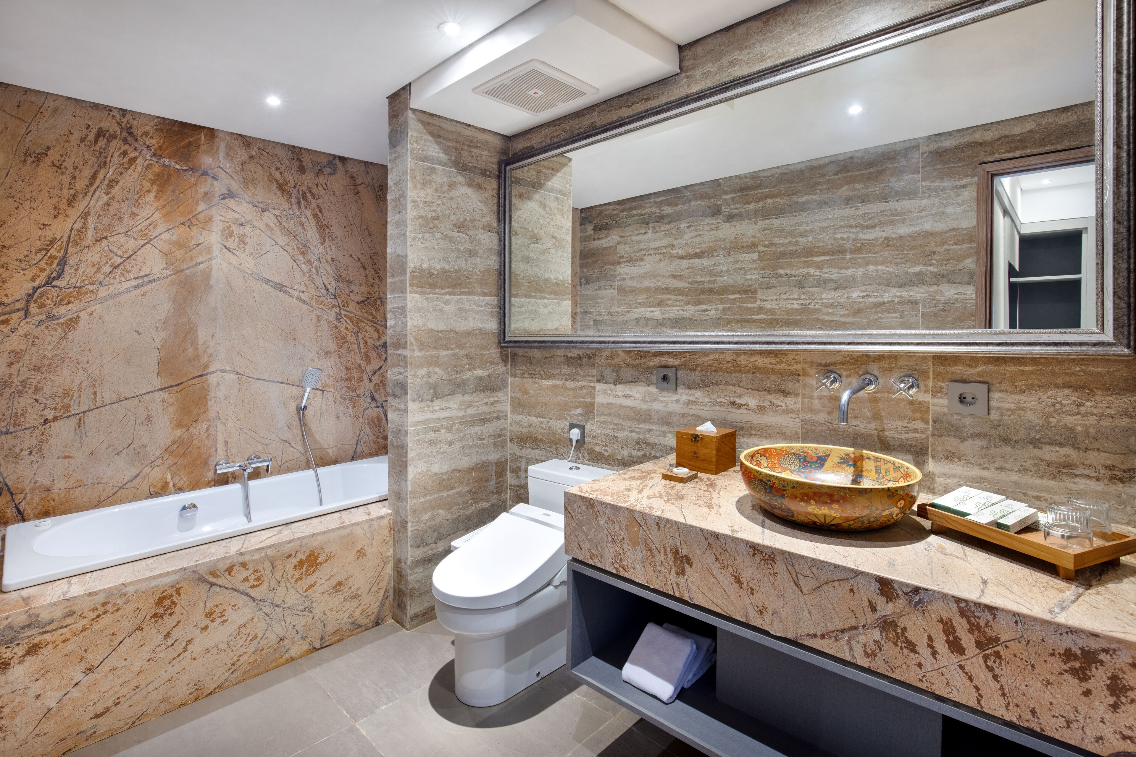 A modern bathroom with a marble wall, a large mirror, a bathtub, a toilet, and a vanity with a bowl sink.