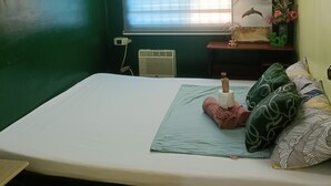 Et værelse med seng, et vindue med persienner, en vægmonteret ventilator, en varmeblæser og et lille bord med dekorative genstande.