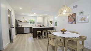 A modern kitchen with a central island, bar stools, and a dining table with a bowl of fruit. - Pet friendly beach house w/private backyard & outdoor shower- steps from beach (St. Augustine)
