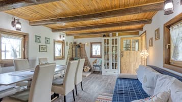 A rustic interior with wooden beams, a dining area with chairs and tables, a blue patterned sofa, and a fireplace.