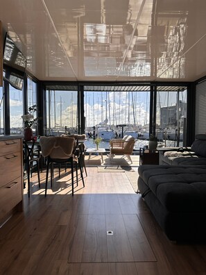 A modern interior with a large window overlooking a marina, wooden flooring, and a seating area with a sofa and chairs.