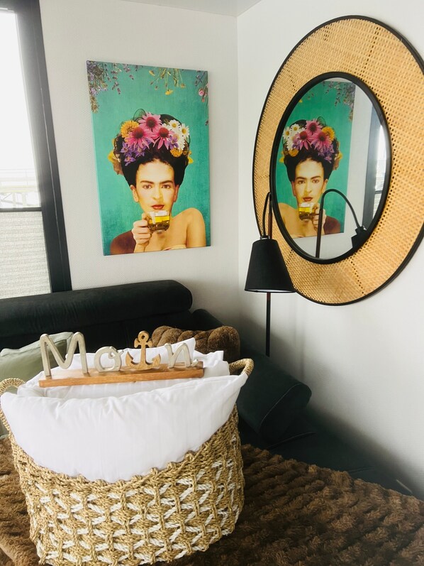 Een woonkamer met een geweven mand, een lijstwerk schilderij, een ronde spiegel, een zwarte bank en een klein houten bord.