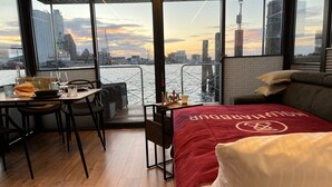 Ein Zimmer mit Blick auf eine Stadtlandschaft, eine Essecke mit Stühlen und einem Tisch sowie ein Bett mit roter Decke.