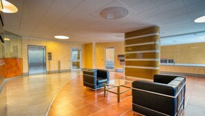 A lobby with a reception desk, two black leather sofas, a glass coffee table, a large pillar, and an elevator. - GuestHost - Cherenel House (Milan)
