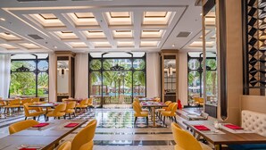 A spacious dining area with multiple tables and yellow chairs, large windows with a view of greenery, and a modern ceiling design.