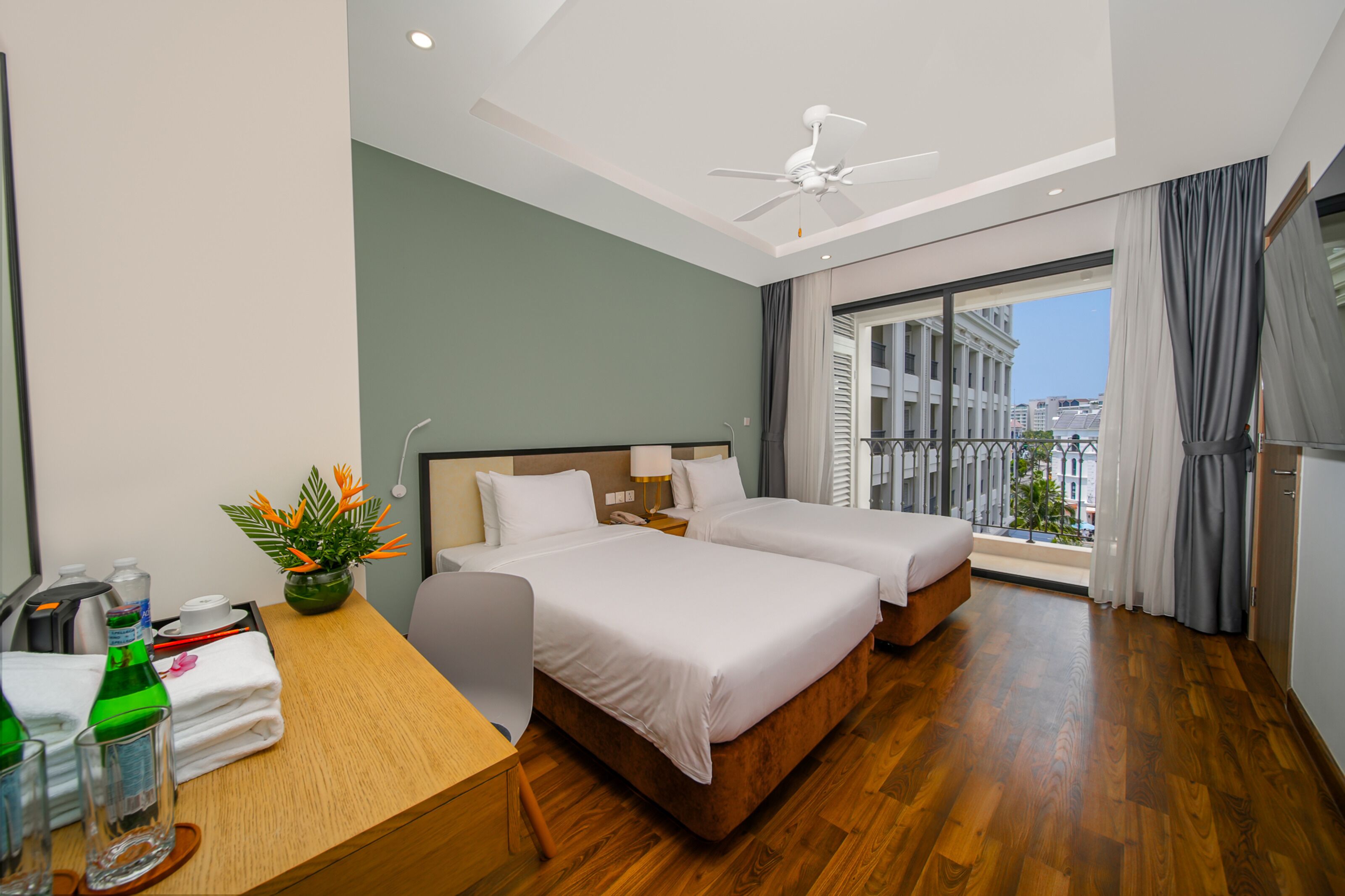 A hotel room with two beds, a desk with toiletries, a potted plant, and a balcony with a city view.
