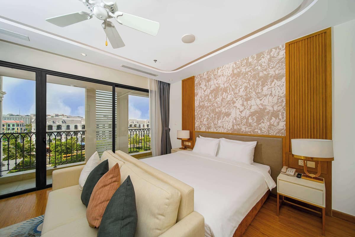 Habitación de hotel con una cama grande, un sofá, un ventilador de techo, una mesita de noche con lámpara y vistas a un paisaje urbano a través de ventanas amplias.