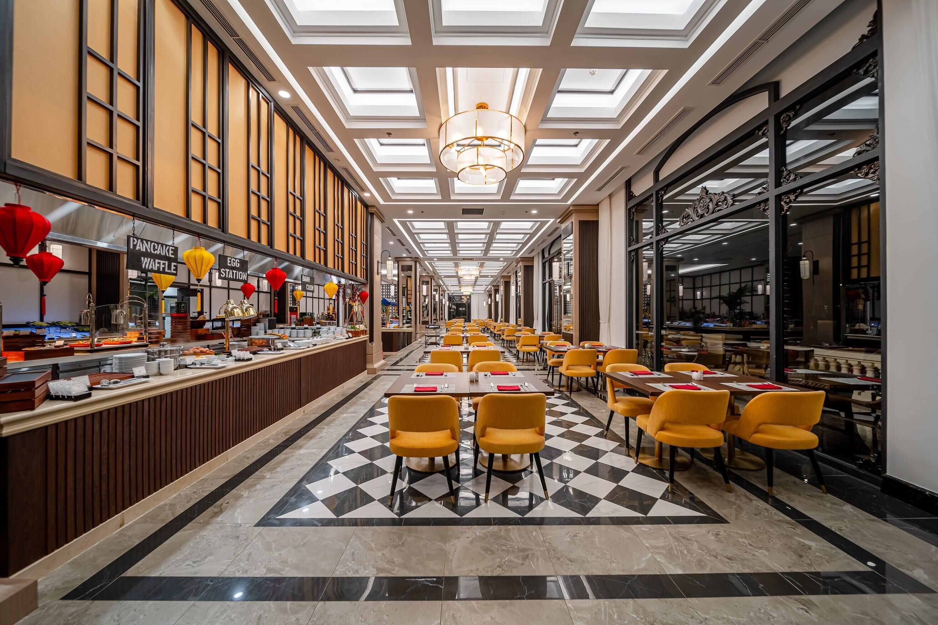 A spacious hotel lobby with a checkered floor, yellow chairs, a long reception desk, and a modern chandelier.