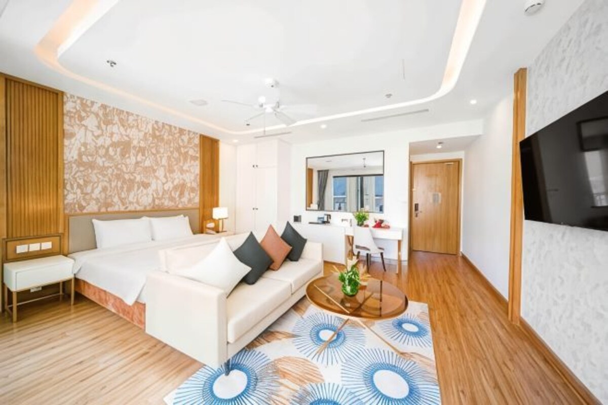 Une chambre d’hôtel moderne dotée d’un grand lit, d’un coin salon avec un canapé et une table basse, d’un bureau avec une chaise et d’un téléviseur fixé au mur.