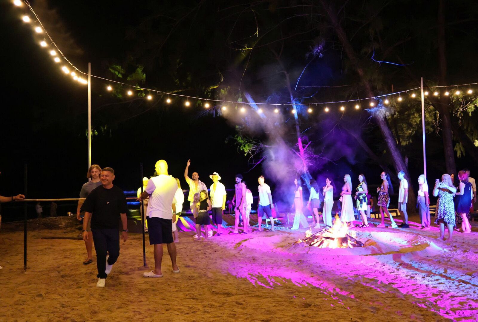 Un grupo de personas reunidas alrededor de una fogata por la noche, con luces colgantes sobre sus cabezas.