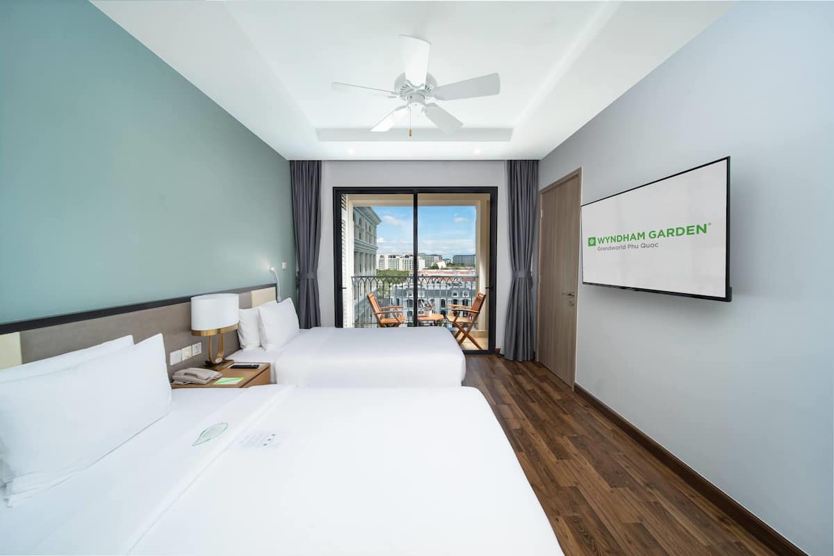 Habitación de hotel con dos camas, ventilador de techo, televisor y balcón con vista a la ciudad.