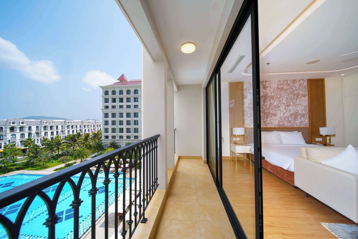 Habitación de hotel con balcón que da a una piscina y vistas del edificio del hotel.