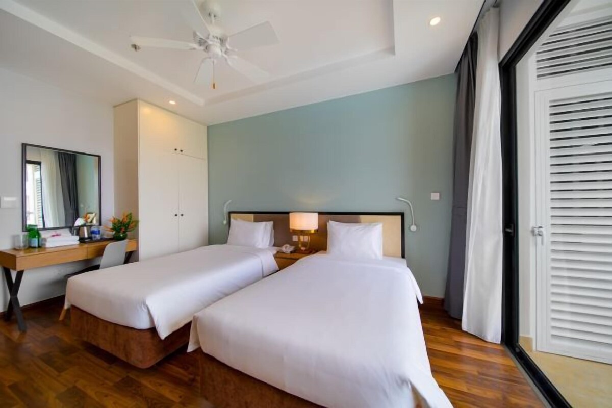 Habitación de hotel con dos camas, un escritorio con silla, un ventilador de techo y un amplio ventanal con persianas.