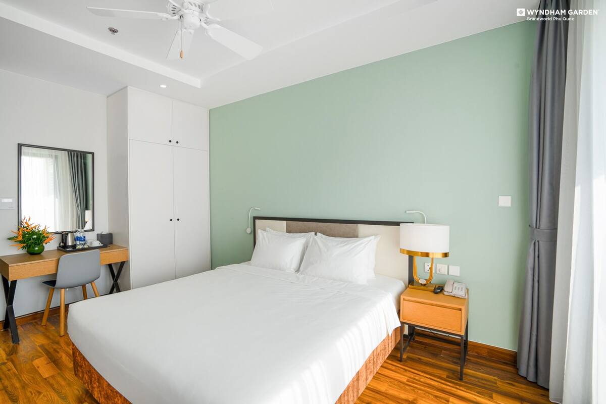 Habitación de hotel con una cama, un escritorio con silla, un armario, una mesita de noche con lámpara y un ventilador de techo.