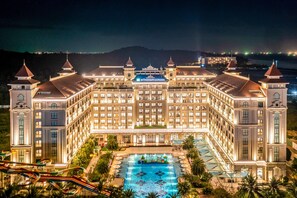 Un hotel grande y bien iluminado con piscina en un patio central, varias alas y montañas al fondo.