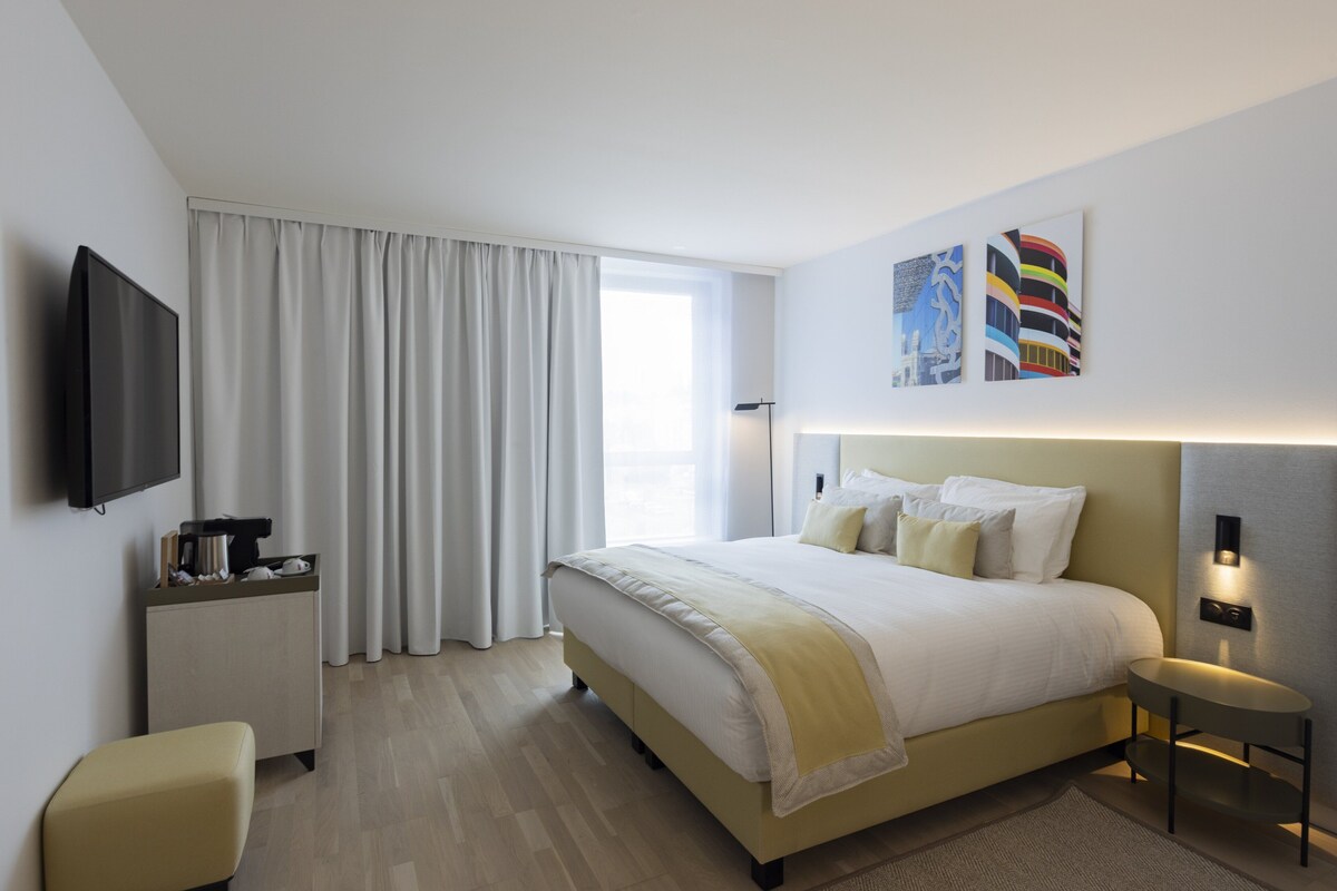 Habitación de hotel moderna con una cama grande, un televisor de pantalla plana, una mesita de noche y un ventanal con cortinas.
