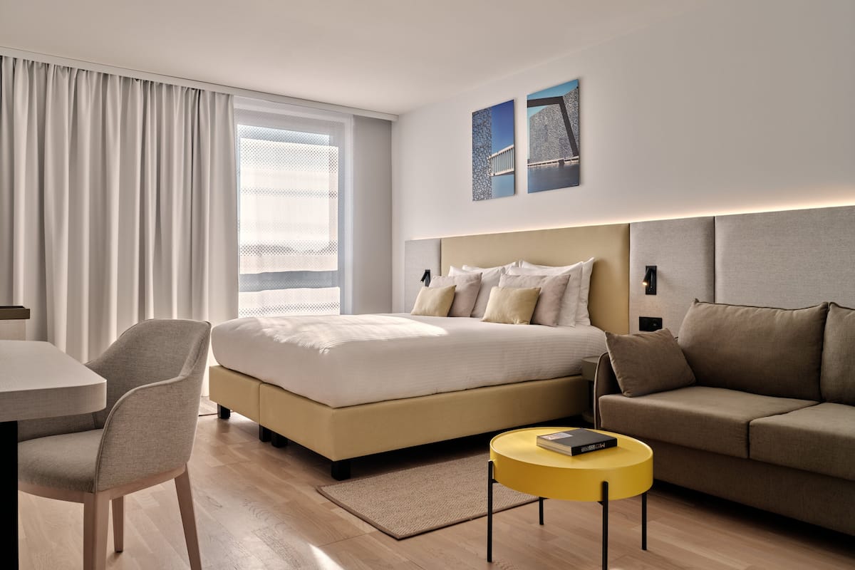 A modern hotel room with a large bed, a sofa, a small round table, and a dining area.