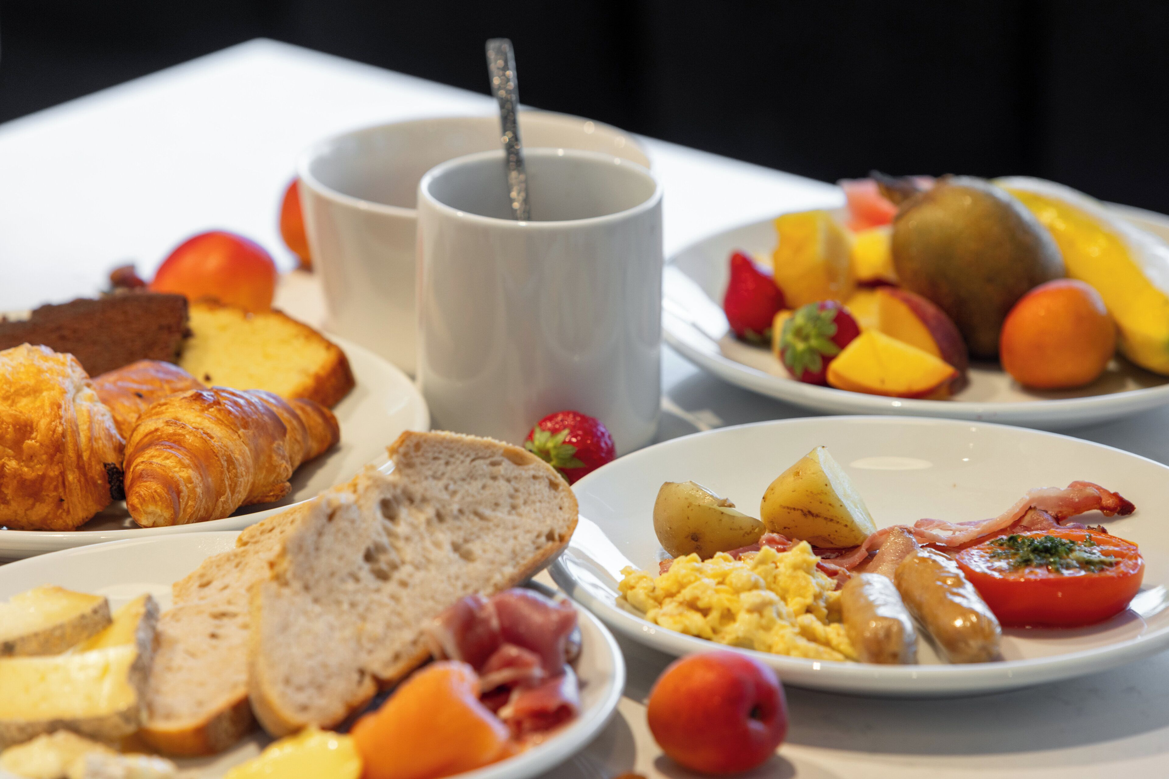 Een ontbijt met croissants, brood, eieren, worstjes, tomaten en fruit op schotels.